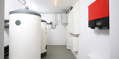 Das Bild zeigt den Pufferspeicher und Trinkwarmwasser-Standspeicher gegenüber von dem Wechselrichter (rot) und dem Doppeltarifzähler.